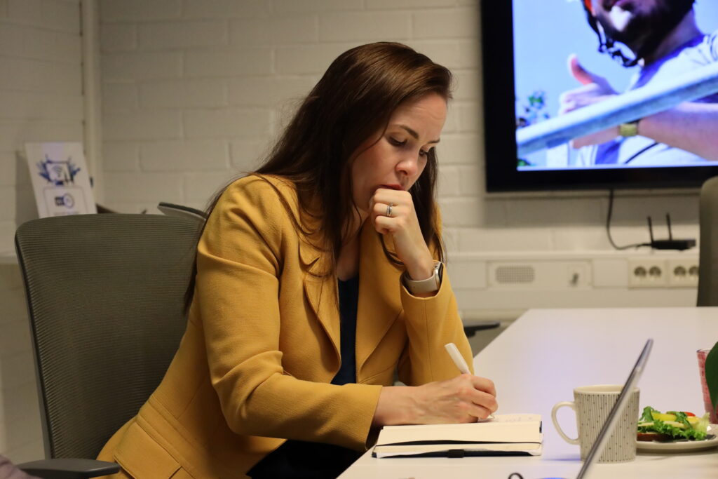 Henkilö istuu neuvotteluhuoneessa valkoisen pöydän ääressä ja kirjoittaa muistivihkoon kynällä. Pöydällä on kannettava tietokone, kahvikuppi ja lautanen, jossa on voileipä. Taustalla näkyy valkoinen tiiliseinä ja suuri näyttö, jossa on kuva henkilöstä ulkona, peukalo pystyssä.