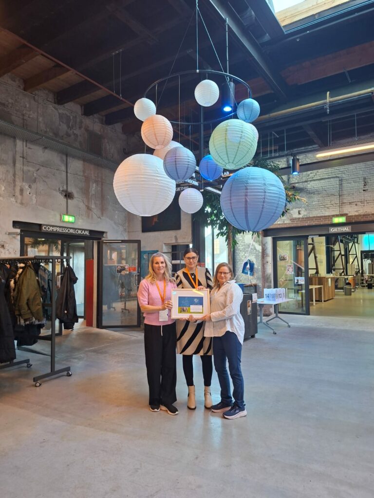 Kolme henkilöä seisoo teollisuustyylisessä sisätilassa ja pitelee yhdessä kehystettyä sertifikaattia tai palkintoa. Yläpuolella roikkuu suuri koristeellinen valaisin, jossa on useita erikokoisia pyöreitä paperilyhtyjä sinisen ja valkoisen sävyissä. Taustalla näkyy lasiovisia tiloja, vaatetelineitä ja kylttejä, joissa lukee muun muassa 'COMPRESSORGRÄL' ja 'GETAN'. Lattia on betonipintainen ja tila on avara.