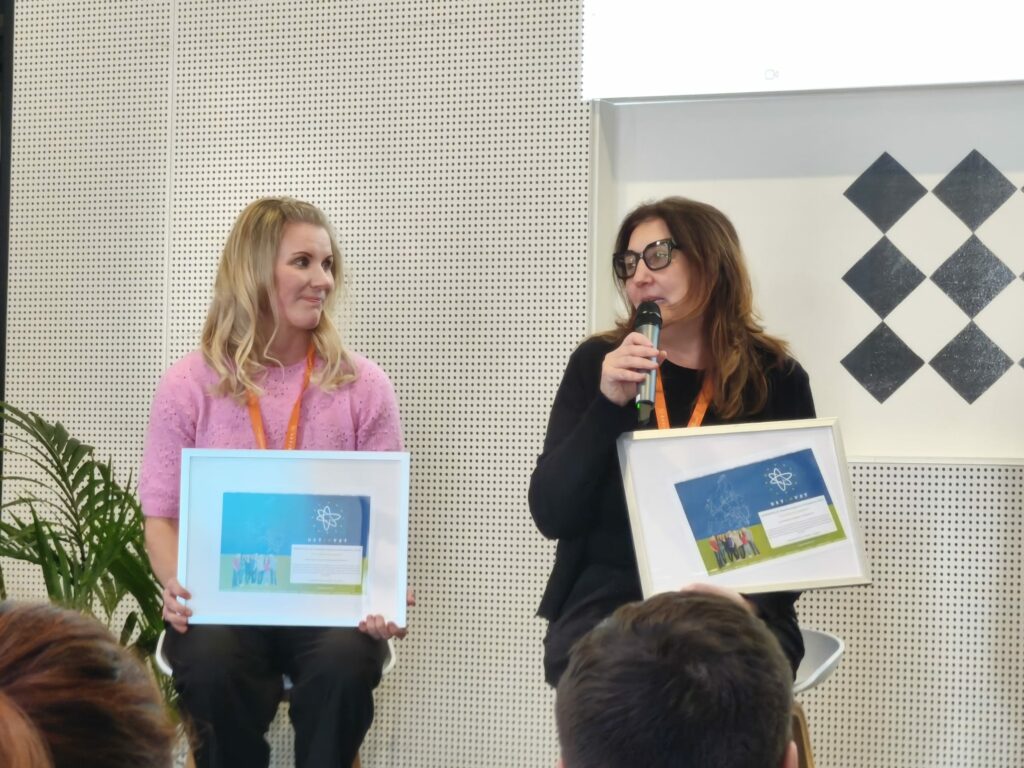 Kaksi henkilöä istuu vierekkäin lavalla tai esiintymisalueella, molemmat pitävät käsissään kehystettyä sertifikaattia tai palkintoa, jossa näkyy sininen ja vihreä tausta sekä NETINVET-logo. Toinen henkilö pitää mikrofonia. Taustalla on vaalea seinä, jossa on mustavalkoinen ruutukuvio ja viherkasvi sivussa. Etualalla näkyy yleisön päitä.