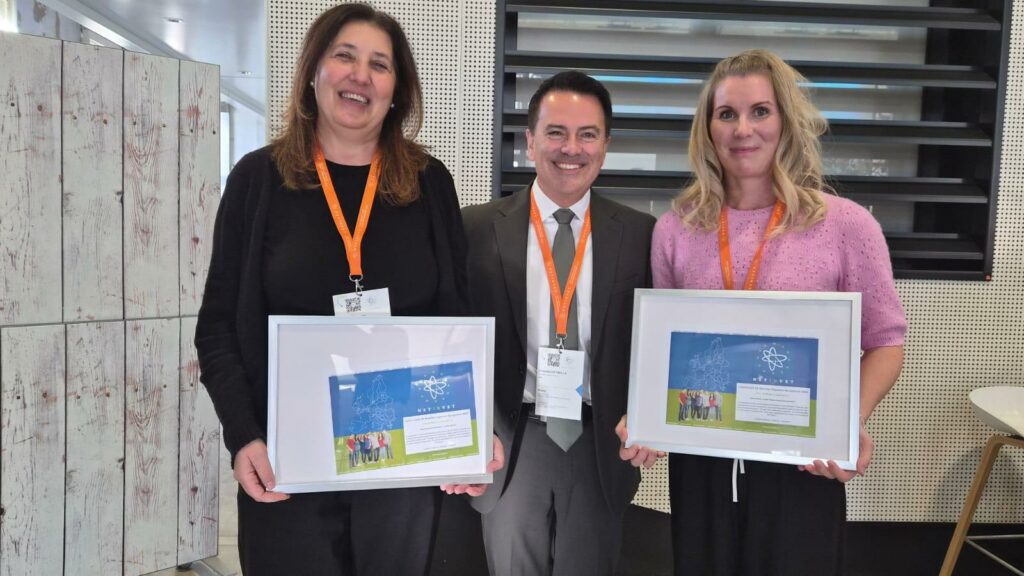 Kolme henkilöä seisoo vierekkäin sisätilassa ja pitää käsissään kehystettyjä sertifikaatteja, joissa näkyy sininen tausta, NETINVET-logo ja ryhmäkuva. Kaikilla on kaulassa oranssit nimikorttinauhat. Taustalla on vaalea seinä, jossa on mustia vaakasäleikköjä, sekä puinen kaappi vasemmalla.