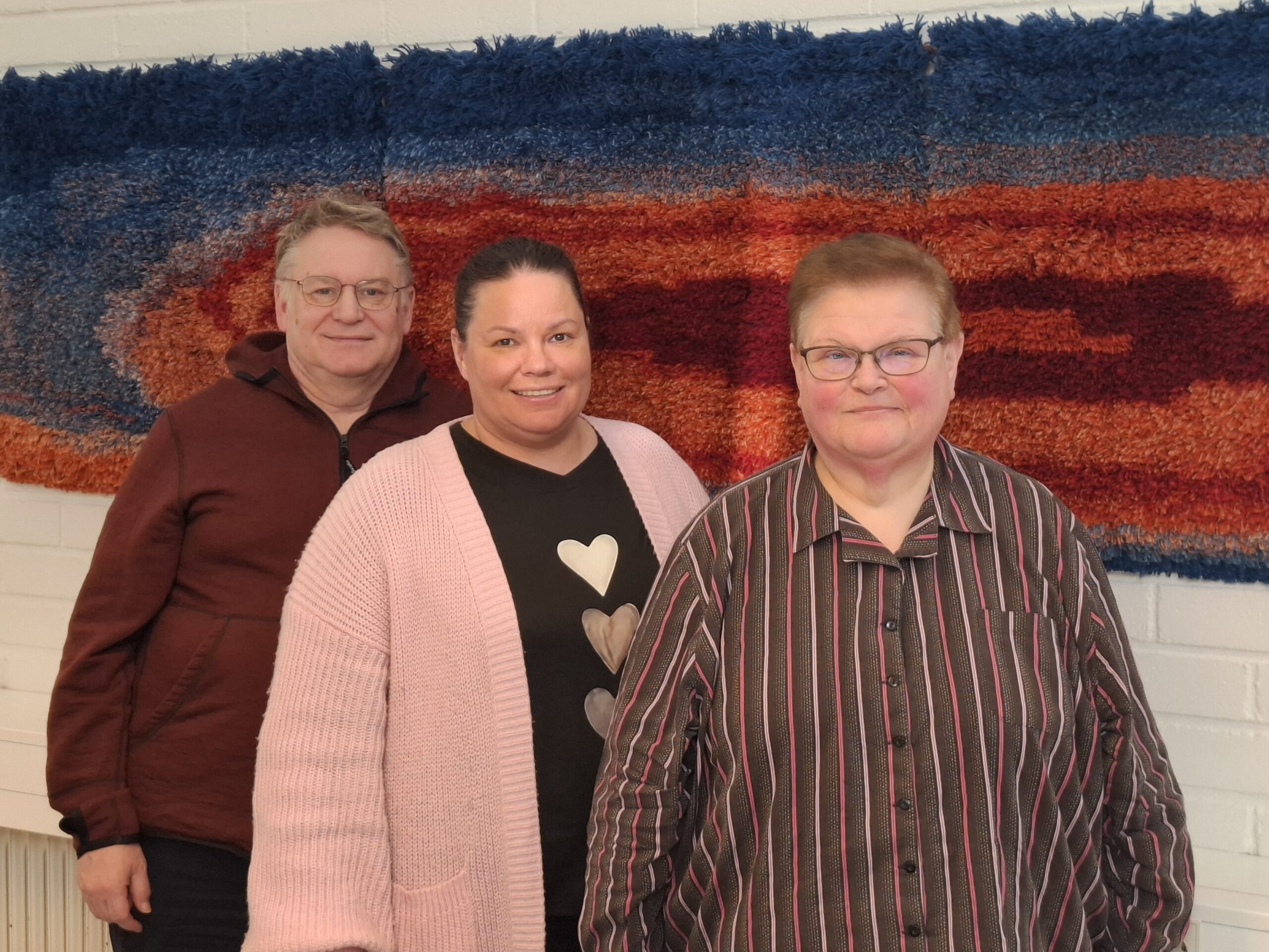 Kuva, jossa kolme henkilö katsoo kameraan kohti. Heidän takanaan on seinällä punasininen ryijy.