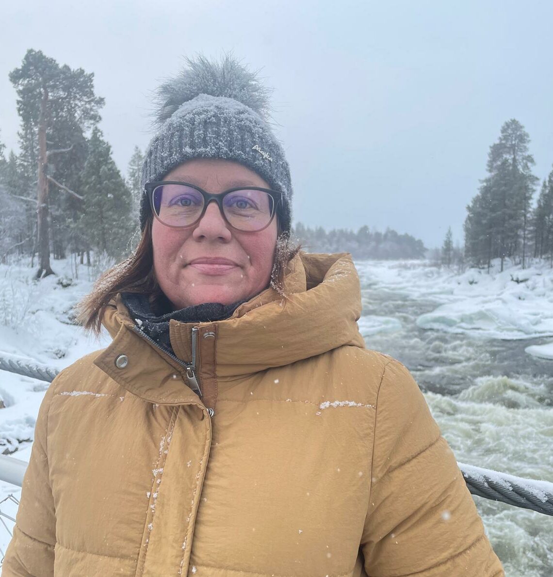 Henkilö katsoo kameraan. Hänen takanaan näkyy talvinen metsä ja koski, joka virtaa.