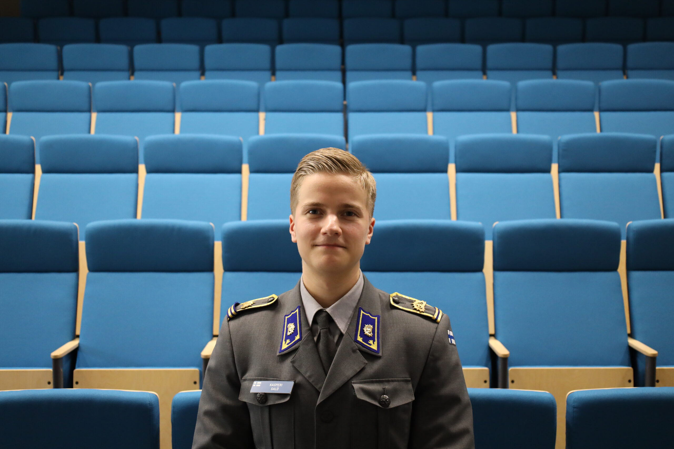 Mies seisoo kameran edessä harmaassa upseerikadetin asussa. Hänen takanaan näkyy monta riviä sinisiä auditorion tuoleja.