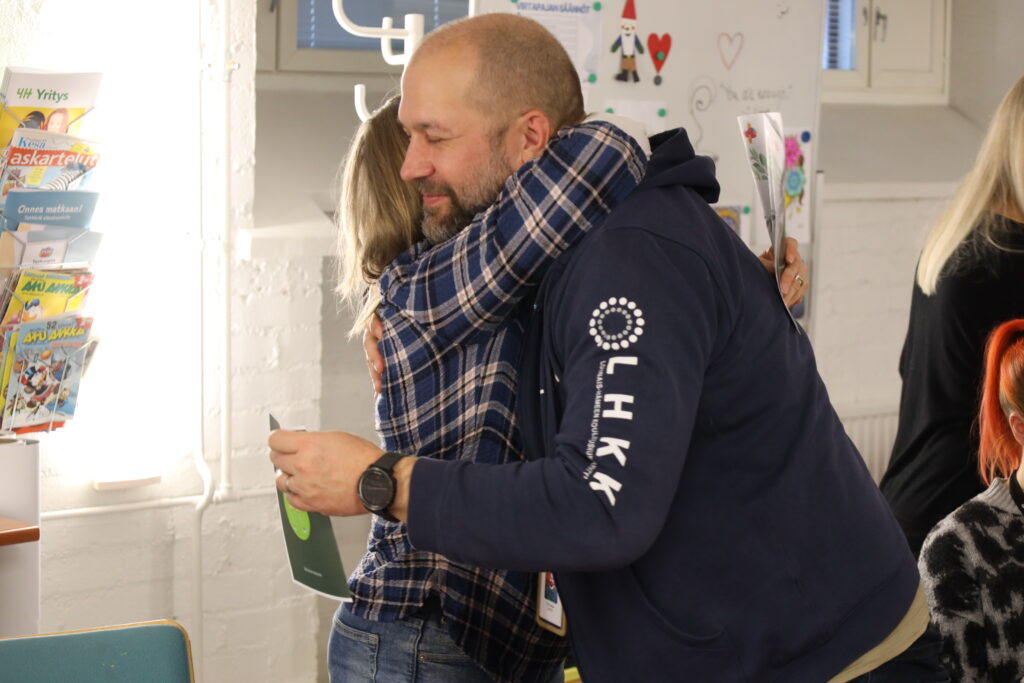 Kaksi ihmistä halaavat. Toisella ihmisellä lukee hihassa LHKK ja siinä on myös LHKK:n logo.