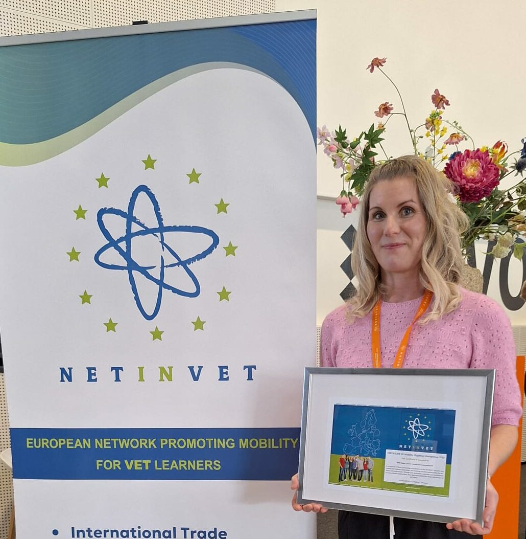 Henkilö seisoo sisätilassa pitäen kehystettyä sertifikaattia tai palkintoa. Vieressä on pystybanneri, jossa lukee 'NETINVET – European Network Promoting Mobility for VET Learners' ja listataan alat: International Trade, Transport & Logistics, Automotive Maintenance, Retail sectors. Taustalla näkyy kukka-asetelma.