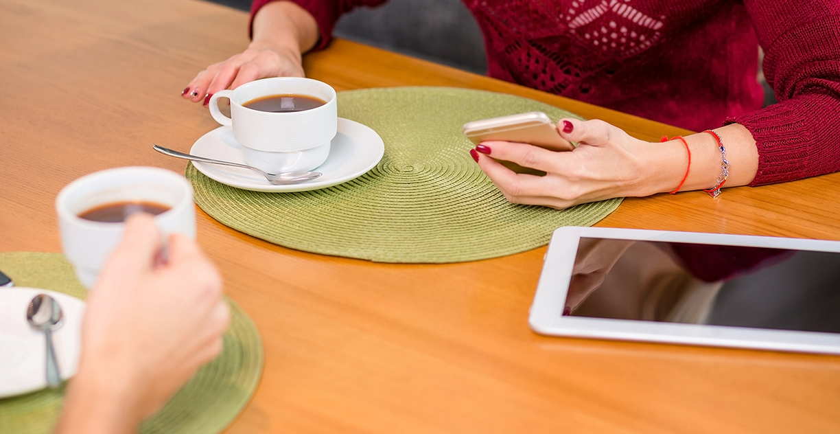 Kuvituskuva, jossa kaksi ihmistä istuu pöydän ääressä juoden kahvia, toisella on kännykkä ja tabletti edessään