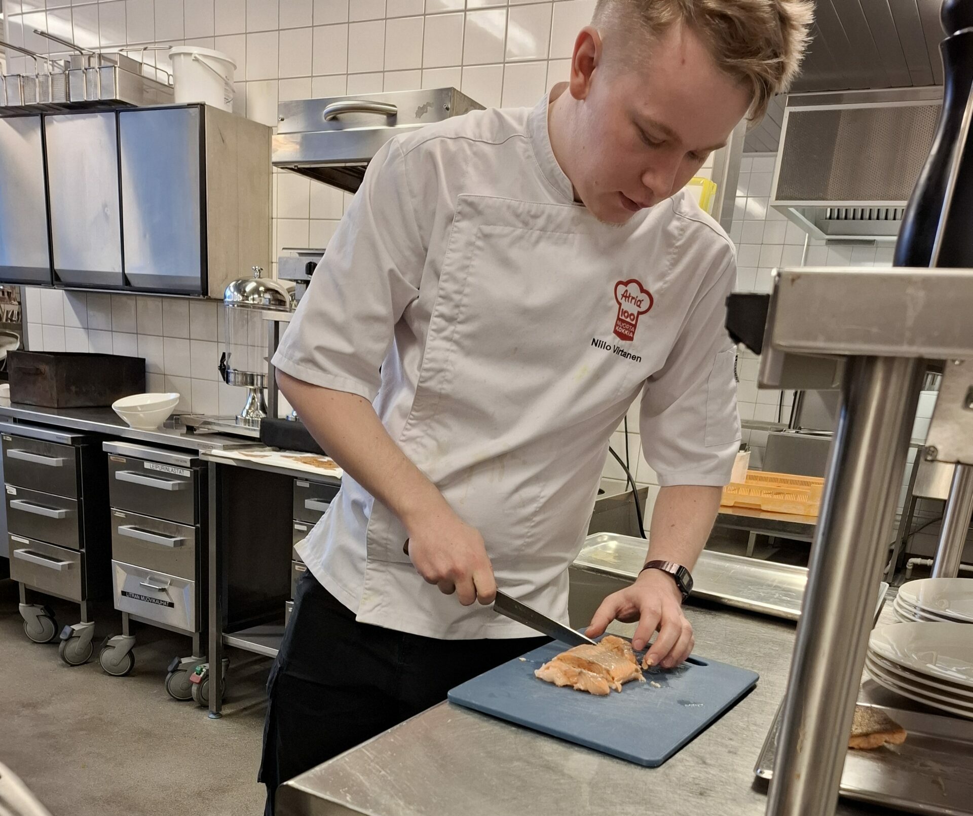 Kokki leikkaa ruokaa leikkuulaudalla. Kokin edessä on paistinpannu, jossa paistetaan kasviksia. Kokilla on valkoinen kokkipaita, ja taustalla näkyy keittiötä.