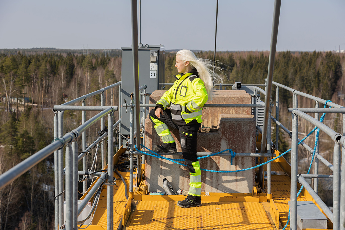 Nainen työvaatteissa seisoo torninosturin päällä ja katselee maisemaa